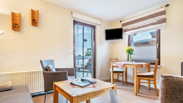 Een moderne woonkamer met een bank, een houten salontafel, een aan de muur bevestigde televisie, een eethoek met een tafel en stoelen, en een schuifdeur die leidt naar een buitenruimte.