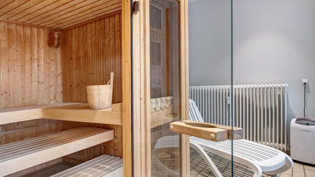 A sauna room with wooden benches, a glass door, a white radiator, and a white chair. - Studio Apartment 'Schwarzwaldliebe' with Pool, Sauna, Mountain View, Balcony & Wi-Fi (Schönwald im Schwarzwald)