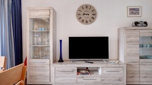 A modern living room with a flat-screen TV, wooden cabinets, a clock, and a framed picture on the wall. - Studio Apartment 'Schwarzwaldliebe' with Pool, Sauna, Mountain View, Balcony & Wi-Fi (Schönwald im Schwarzwald)