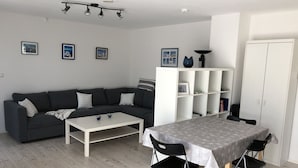 Un salon moderne avec un canapé gris, une table basse blanche, une table à manger recouverte d’une nappe et une étagère blanche.