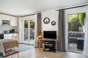 Una sala moderna con un televisor de pantalla plana, una mesa de comedor con sillas, una zona de cocina con electrodomésticos, y vistas a una pared de piedra y follaje exterior.