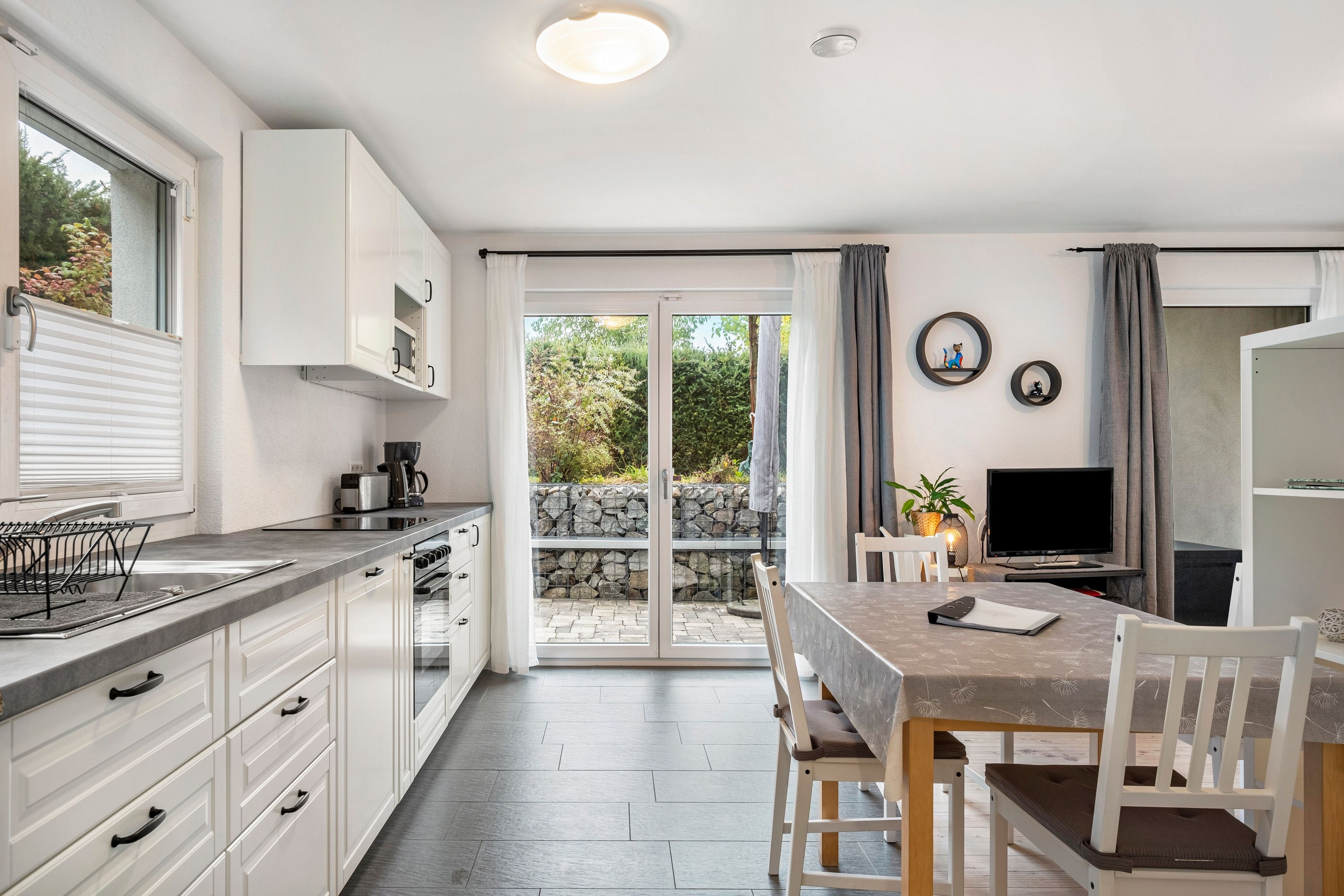 Cozinha moderna com armários brancos, área de jantar com mesa e cadeiras, e porta de vidro deslizante para um jardim.