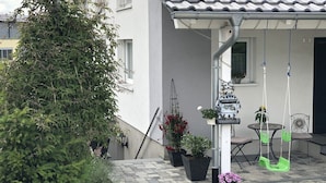 Un patio couvert avec un toit en tuiles, des plantes en pot, une balançoire et une boîte aux lettres.