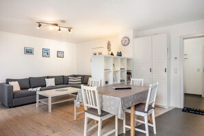 A modern living room with a grey sofa, a white coffee table, and a dining table with white chairs. There are bookshelves and a clock on the wall.