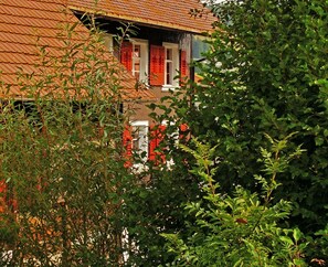 A building with a red-tiled roof, a window with red shutters, and dense green foliage in the foreground. - Holiday Home 'Bernau' with Mountain View, Private Terrace and Wi-Fi (Bernau im Schwarzwald)