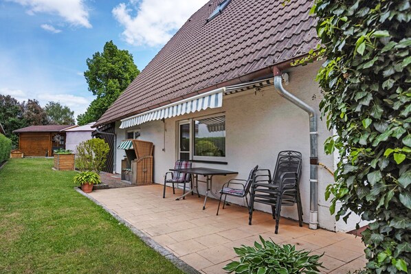 Une maison avec un toit en tuiles, un patio couvert avec des chaises et une table, et un jardin agrémenté de plantes en pot.