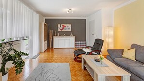 A modern living room with a sofa, a coffee table, and a chair. There is a kitchen area in the background with a cabinet and a painting on the wall.