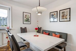 Un coin repas avec une table blanche, des chaises noires, un banc avec des coussins rouges et des œuvres d’art encadrées accrochées aux murs.