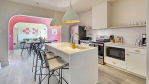 A modern kitchen with a central island, bar stools, a pink accent wall, and a dining area with a pink sofa.