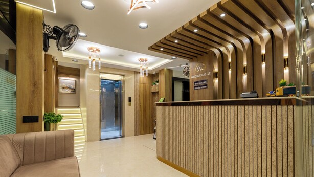 A modern reception area with a wooden reception desk, a seating area with a sofa, and a hallway leading to an elevator. - Hotel Windsor Castle (Mumbai)