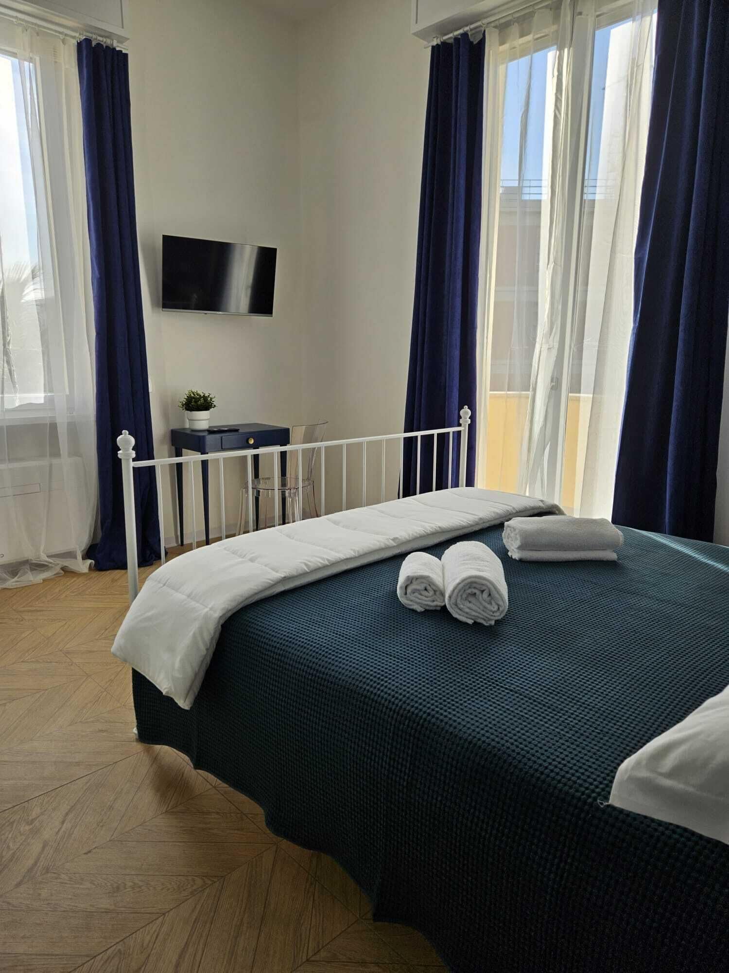 Una cama bien hecha con sábanas blancas y toallas enrolladas, una mesita de noche con una planta y un televisor en la pared.