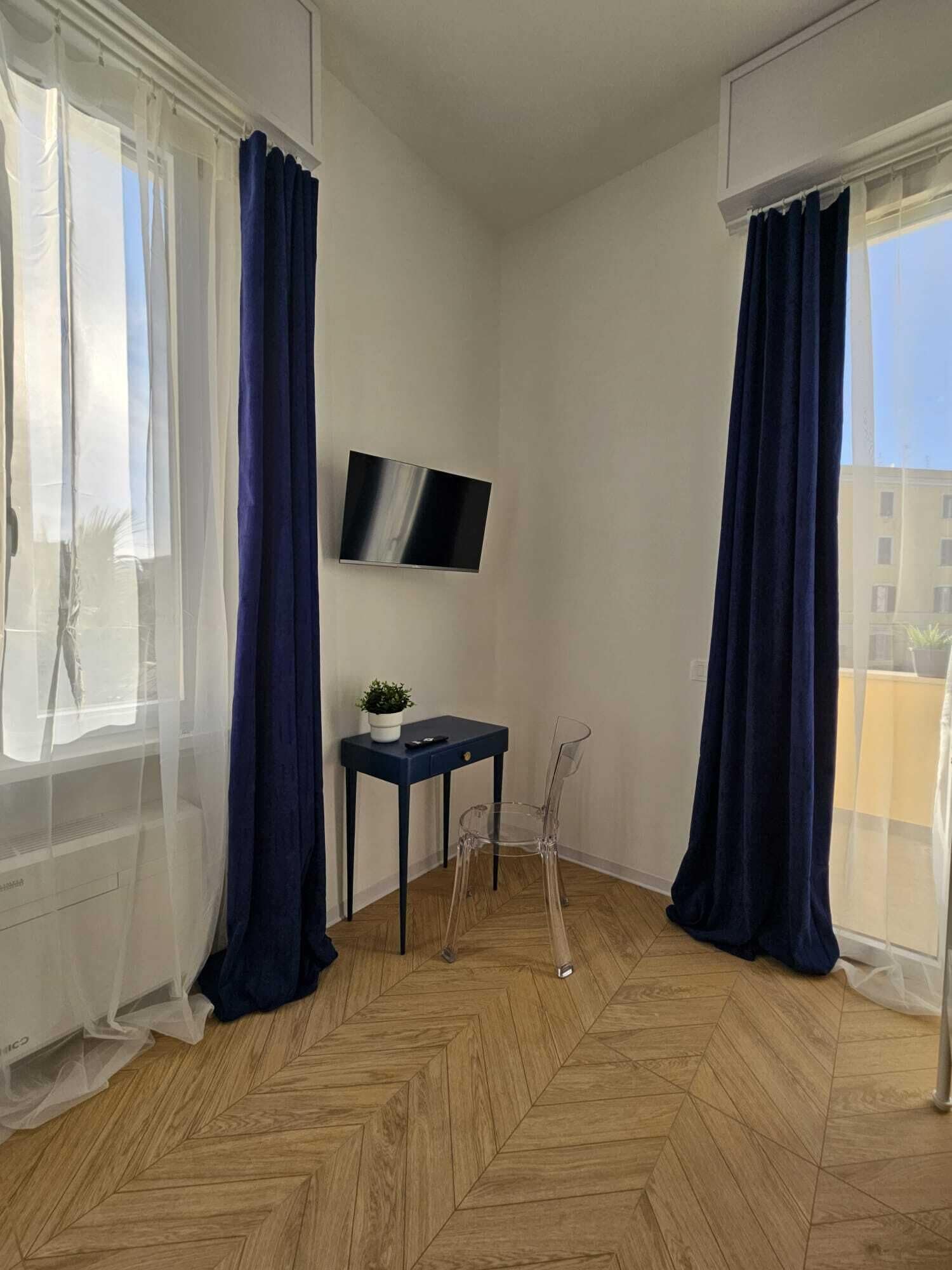 Quarto com piso de madeira em padrão losango, uma pequena mesa com uma planta, uma cadeira e TV na parede.