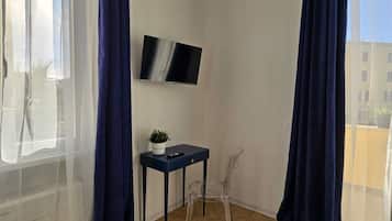 Quarto com piso de madeira em padrão losango, uma pequena mesa com uma planta, uma cadeira e TV na parede.