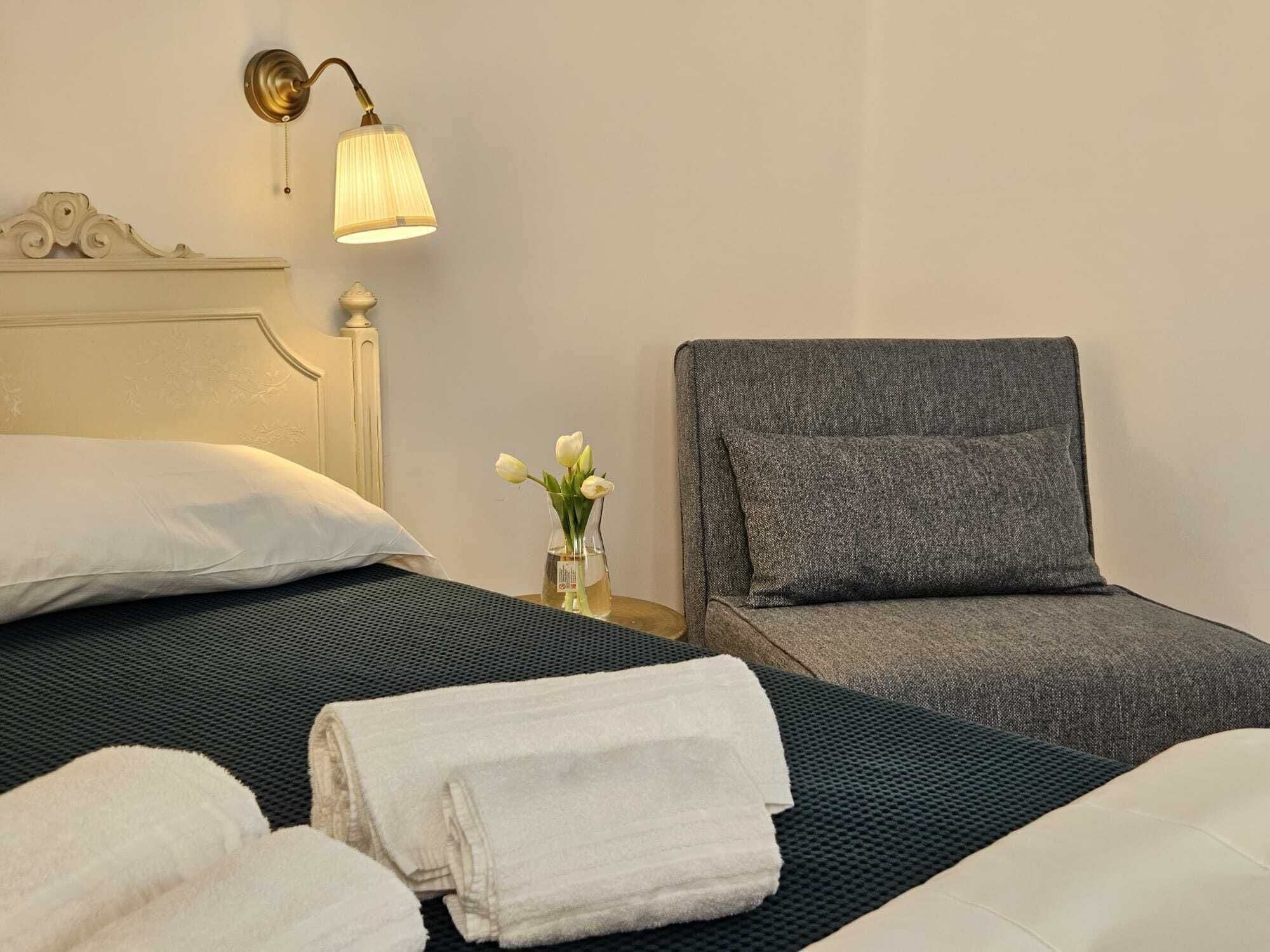 Una habitación de hotel con una cama, un sillón gris y una mesita de noche con una lámpara y un florero.