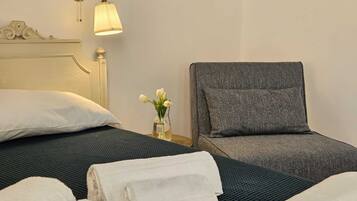Una habitación de hotel con una cama, un sillón gris y una mesita de noche con una lámpara y un florero.
