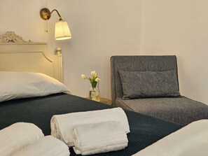 A hotel room with a bed, a grey armchair, and a nightstand with a lamp and a vase of flowers. - Guest House Vanvitelli (Civitavecchia)