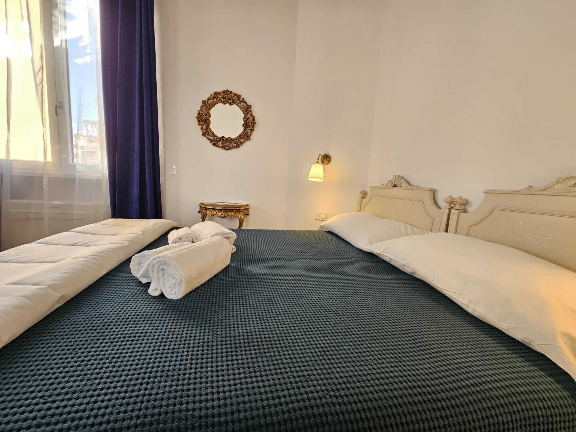 Quarto de hotel com duas camas, um espelho redondo, uma mesinha e uma luminária na parede.