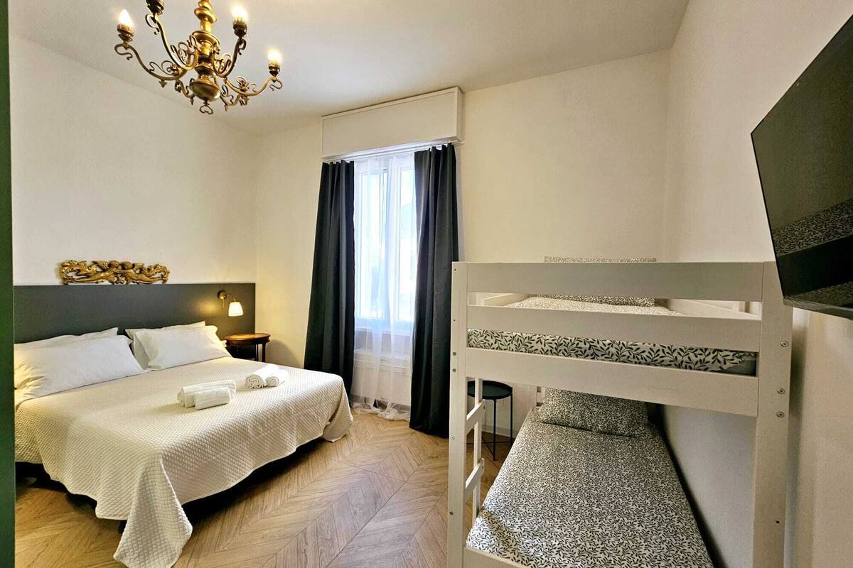 A hotel room with a bed, a bunk bed, a chandelier, and a window with curtains.