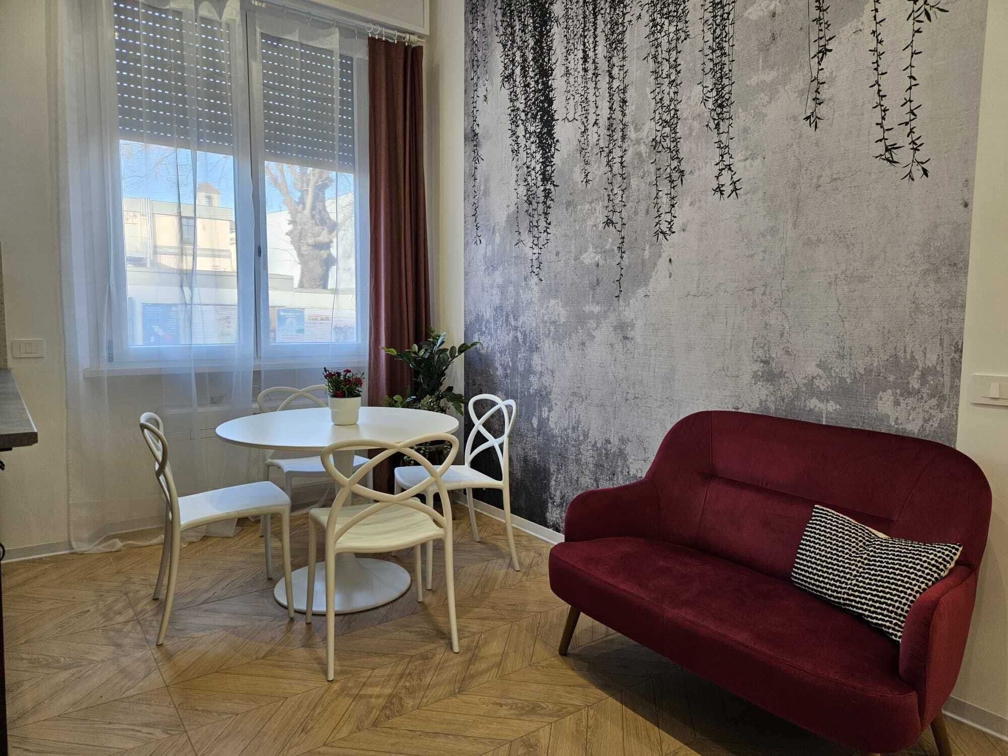 Un comedor con una mesa redonda, cuatro sillas, un sofá rojo y una pared decorada con plantas colgantes.