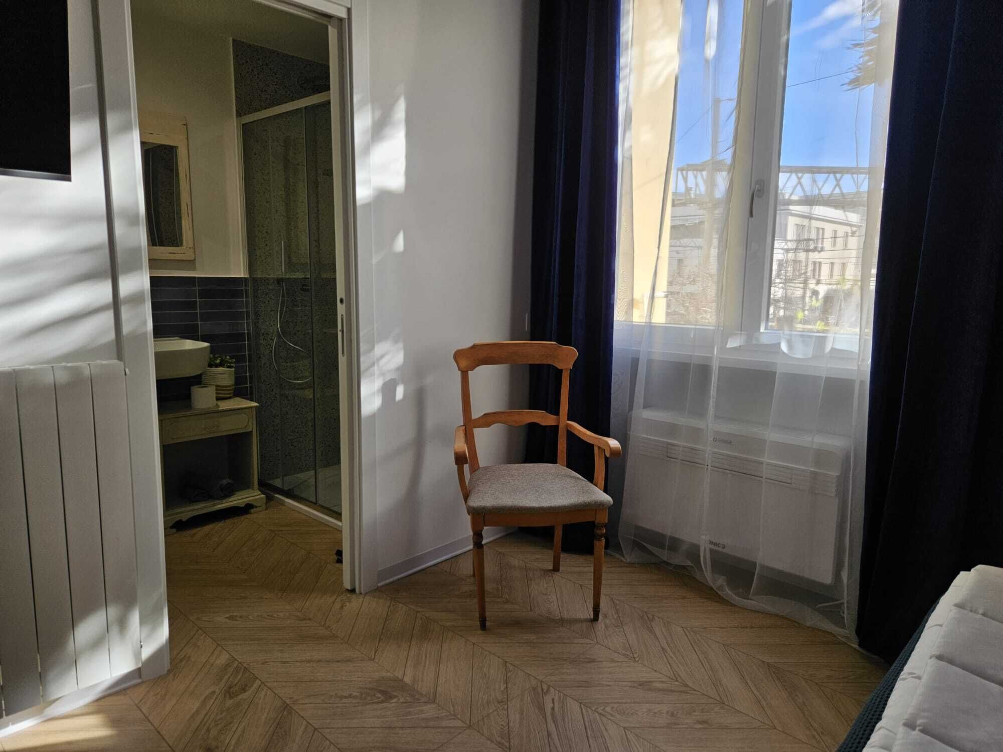 Una habitación con una silla de madera, vista a un edificio a través de una ventana y un baño visible por una puerta abierta.