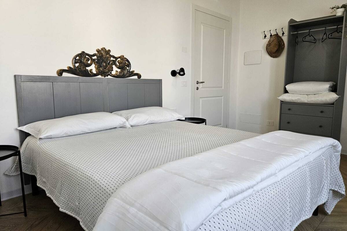 A bedroom with a grey headboard, a bed with white linens, a small bedside table, a grey dresser, and a wall-mounted coat rack.