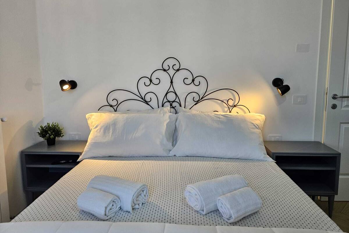 A neatly made bed with white linens and rolled towels, a decorative metal headboard, and two bedside tables with wall-mounted lamps.