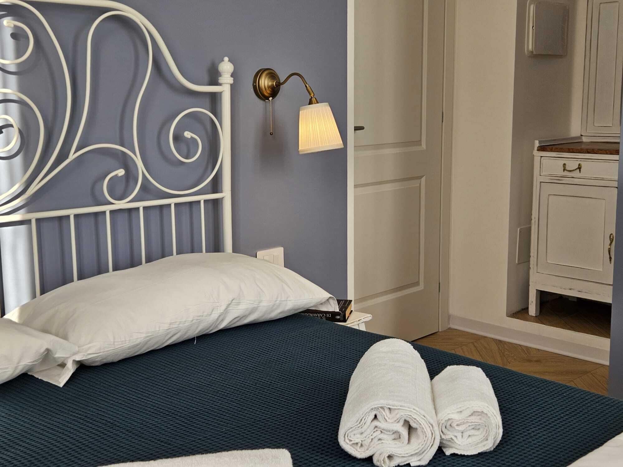 A neatly made bed with white pillows and towels, a white metal headboard, a wall-mounted lamp, and a small white cabinet.