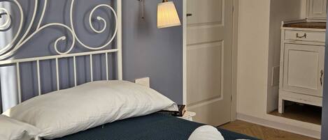 A neatly made bed with white pillows and towels, a white metal headboard, a wall-mounted lamp, and a small white cabinet.