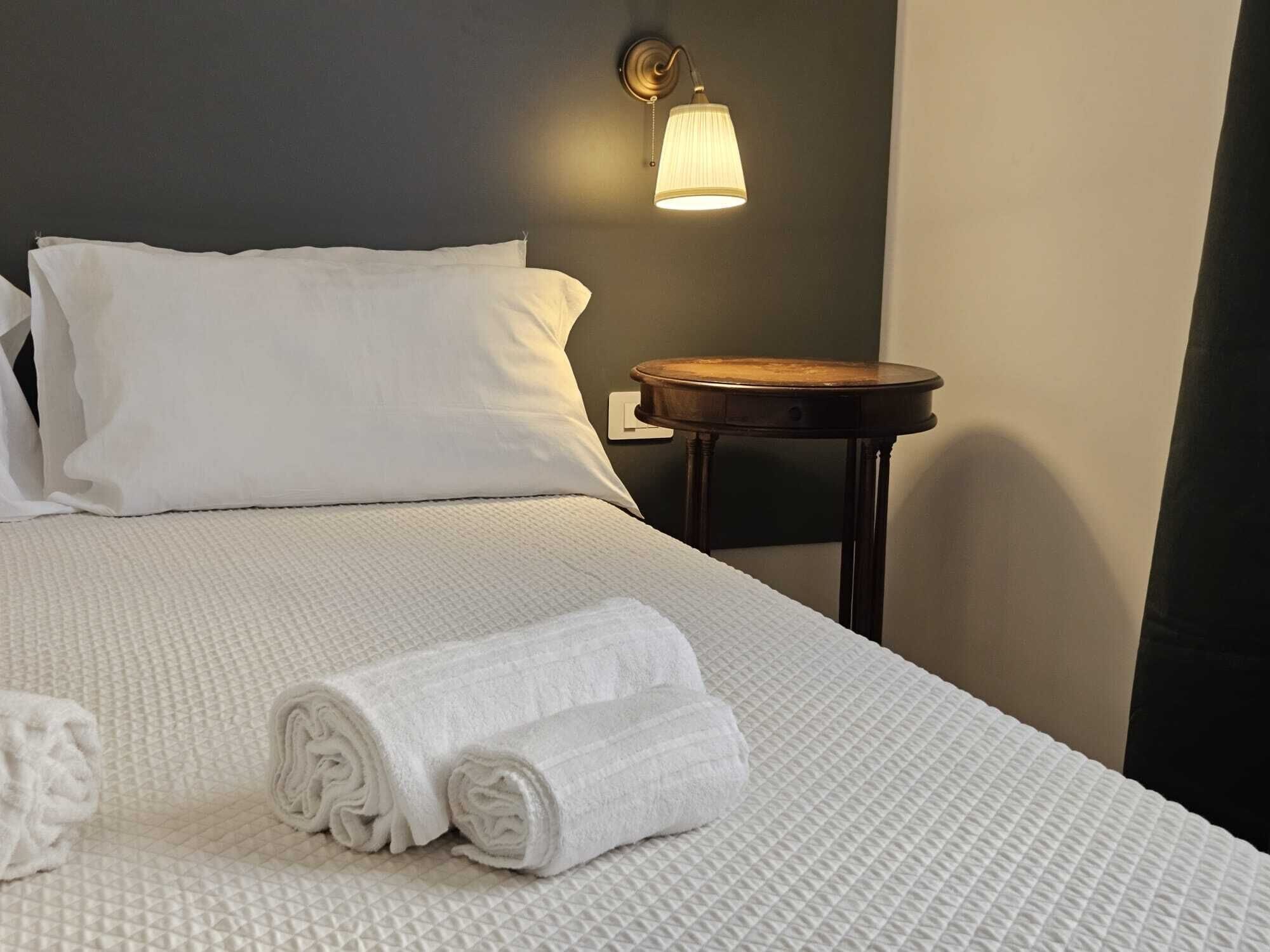 Una cama bien tendida con sábanas y almohadas blancas, una mesita de noche de madera con una lámpara, y toallas enrolladas sobre la cama.