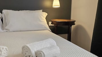 Una cama bien tendida con sábanas y almohadas blancas, una mesita de noche de madera con una lámpara, y toallas enrolladas sobre la cama.