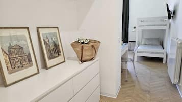 Um quarto com cômoda branca, duas imagens emolduradas, uma cesta de vime e uma cama beliche, além de um radiador.