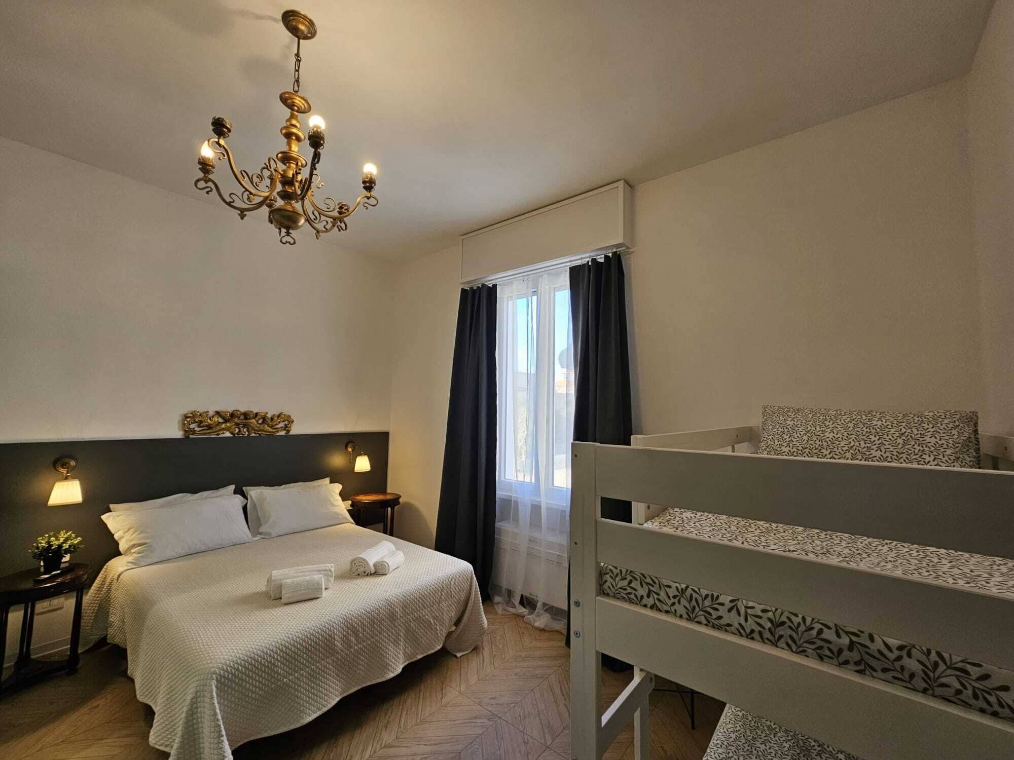 Habitación de hotel con cama, mesita de noche, candelabro, ventana con cortinas y literas.