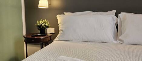 A neatly made bed with white bedding, a bedside lamp, a small table with a plant, and a rolled-up towel on the bed.