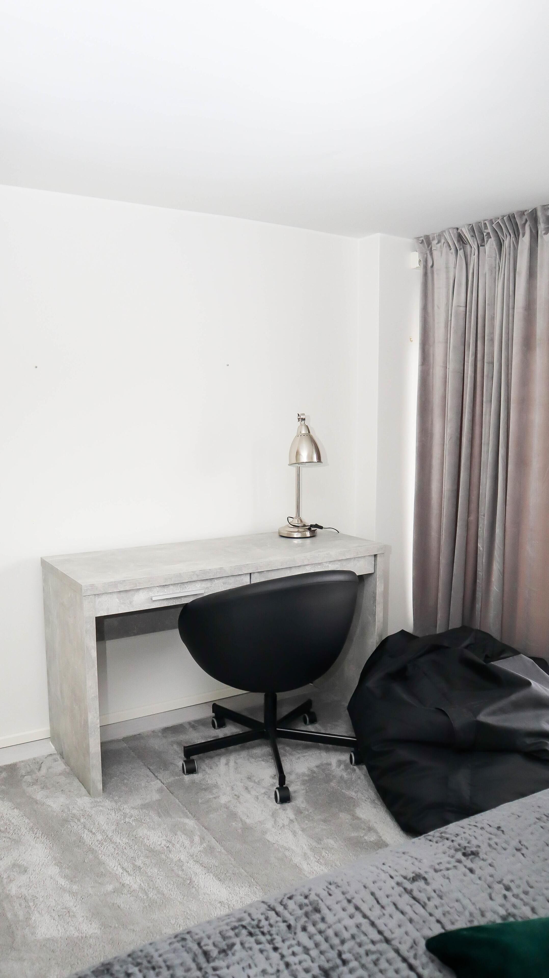 Una habitación minimalista con un escritorio gris, una silla de oficina negra, una lámpara de escritorio y una cortina gris.