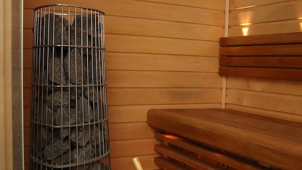 A sauna room with a metal container filled with rocks, a black bucket with a wooden handle, and wooden benches. - Kotimaailma Penthouse Studio (Lahti)