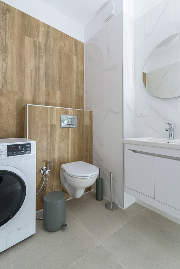A modern bathroom with a wooden wall, a white toilet, a washing machine, and a sink with a mirror. - HUBIX - City of Mara Apartments (Timisoara)