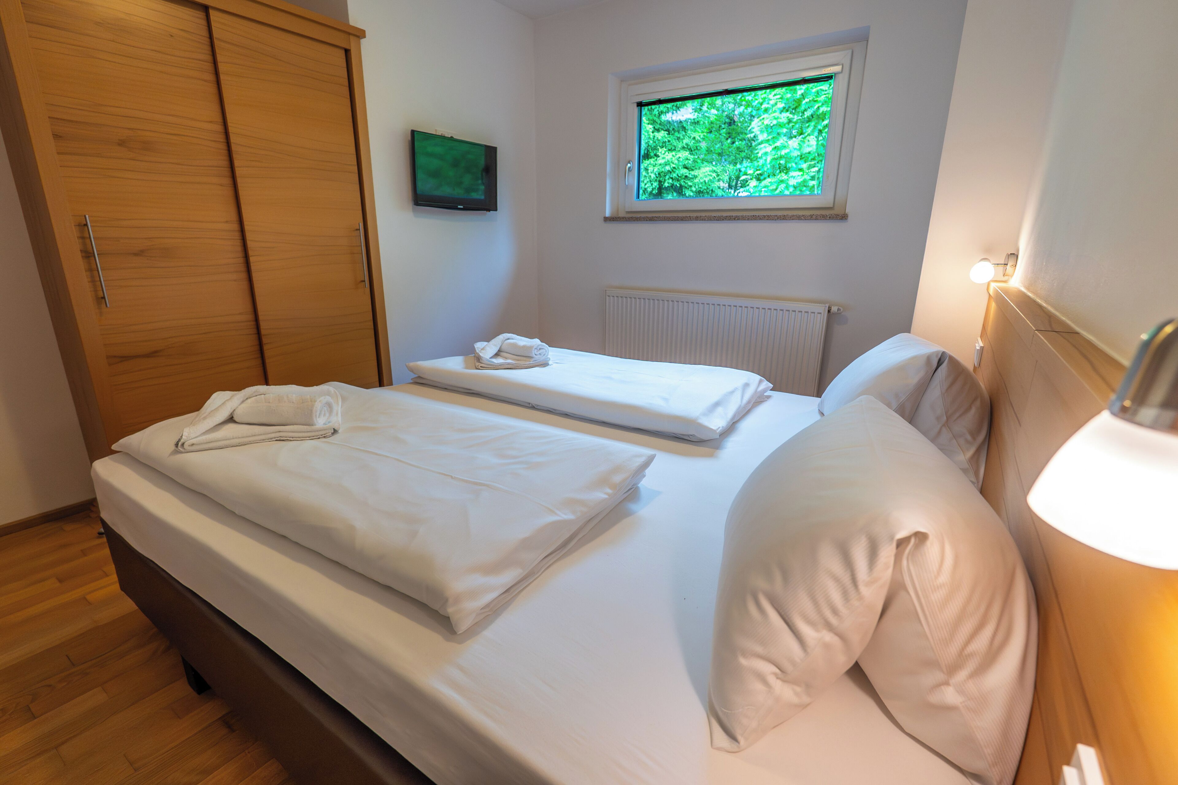 Un dormitorio con dos camas, un armario de madera, una ventana con vistas a zonas verdes y un televisor instalado en la pared.