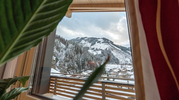 A view from a window with a wooden balcony overlooking a snow-covered mountain and a village. - Appartements MaLo by we rent (Viehhofen)