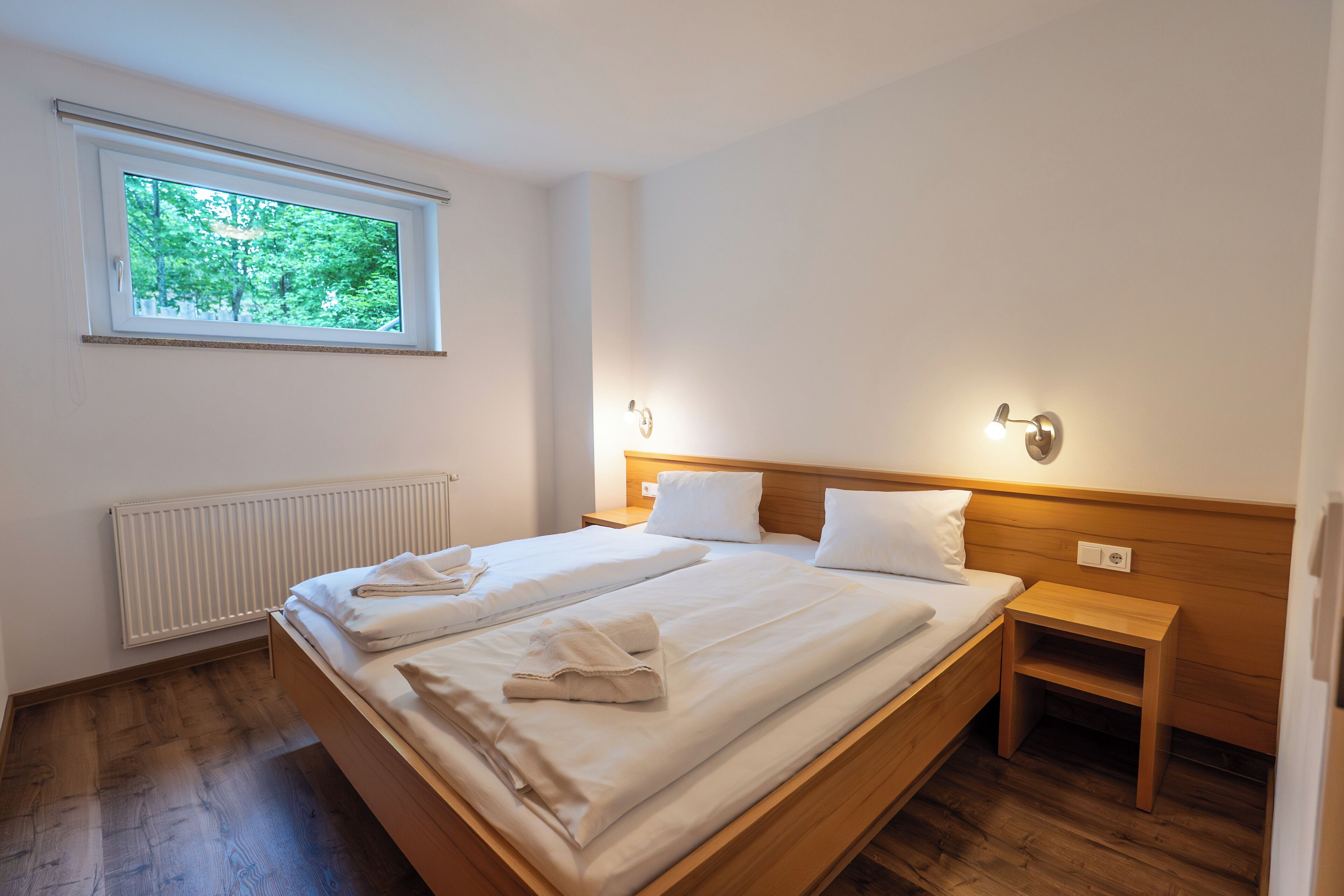 A bedroom with two beds, a window, a bedside table, and a wall-mounted light fixture.