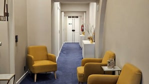 En hotellkorridor med ett blått golv, två gula fåtöljer, ett litet bord med en vas och en vit dörr längst ner.