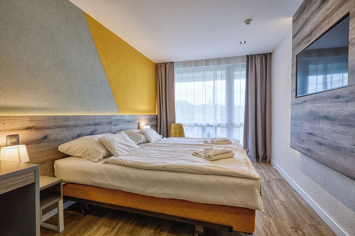Quarto de hotel moderno com uma cama grande, mesa de cabeceira e vista para a cidade através de amplas janelas.
