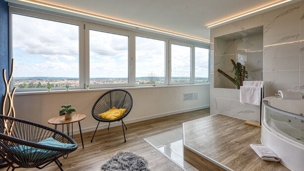A modern room with large windows, a wooden floor, a round rug, a chair, a small table with a plant, and a bathroom with a bathtub. - Hotel Szieszta (Sopron)