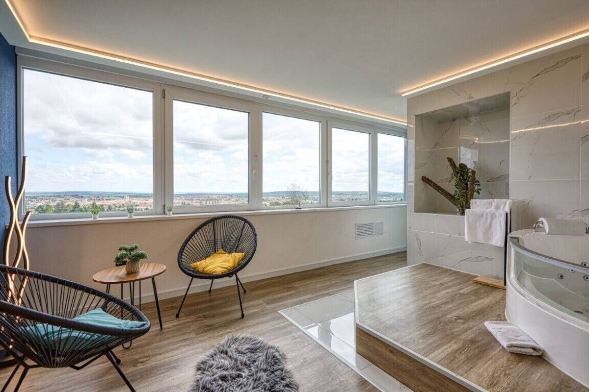 Quarto moderno com amplas janelas, piso de madeira, tapete redondo, uma cadeira, uma mesinha com uma planta e um banheiro com banheira.