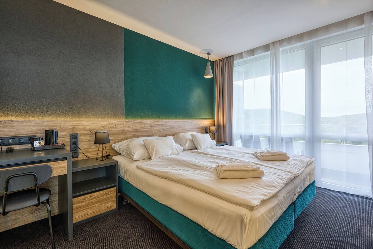 Quarto de hotel com cama, escrivaninha, cadeira, abajur e uma janela grande com cortinas.