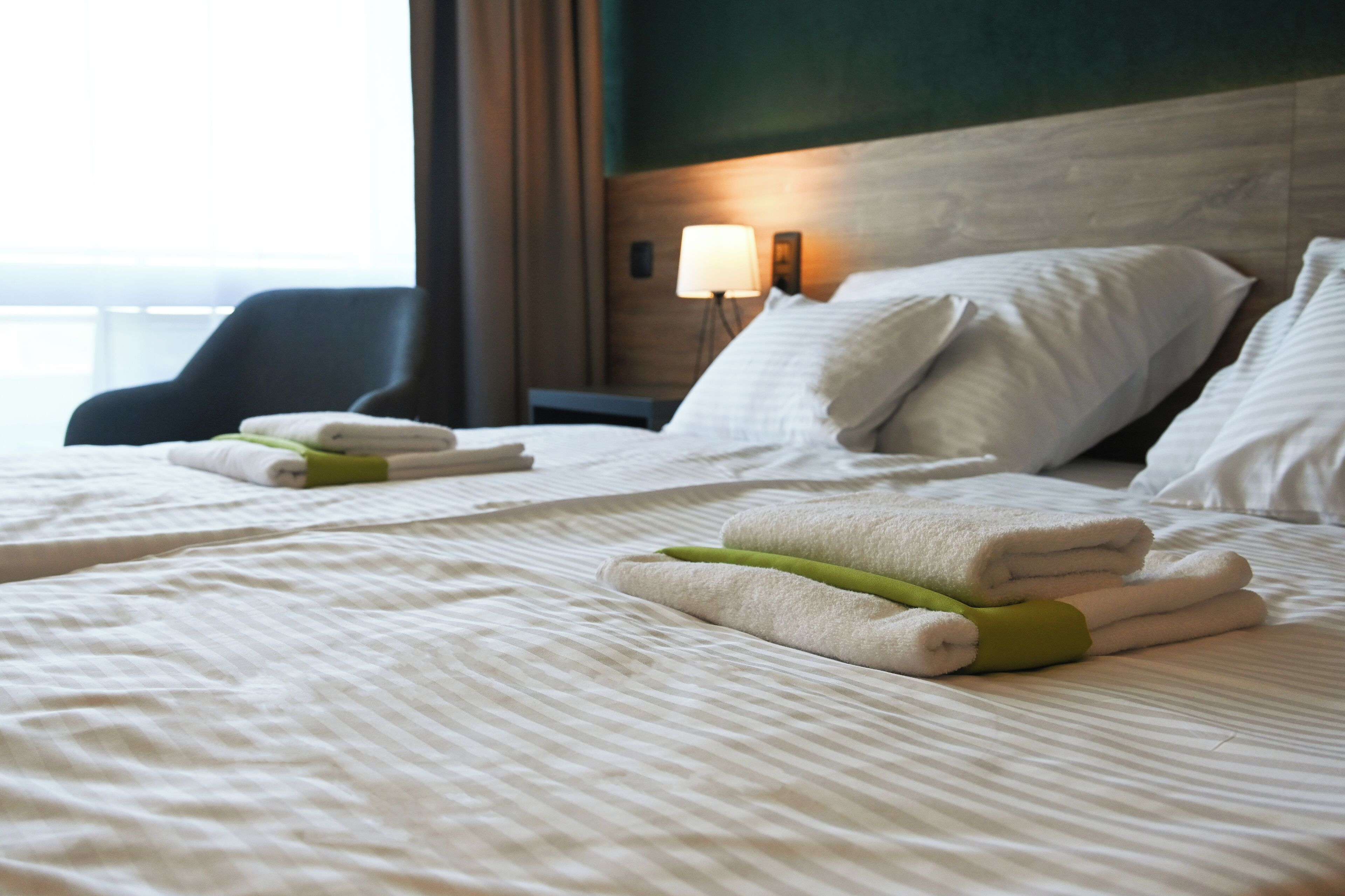 Habitación de hotel con una cama bien hecha, una mesita de noche con lámpara y vistas al exterior por la ventana.