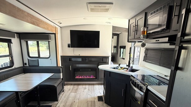 A compact RV kitchen with a flat-screen TV, a built-in fireplace, and a dining area with a bench. - Toy Hauler Vengence Rouge (Brentwood)