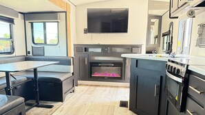 A compact kitchen with a built-in fireplace, a dining area with a table and bench, and a kitchenette with a stove and refrigerator. - Toy Hauler Vengence Rouge (Brentwood)