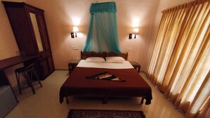 A hotel room with a bed, a desk, a chair, a nightstand, a lamp, and a window with curtains. - HOTEL COCO BECH KOVALAM AYURVEDIC RESORT (Kovalam)