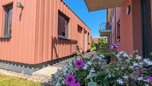 A red building with a window and a door, a balcony, and a garden with flowers.