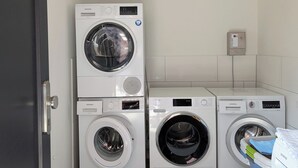 A laundry room with two stacked front-loading washing machines, a white bucket, and a plastic container with laundry detergent.